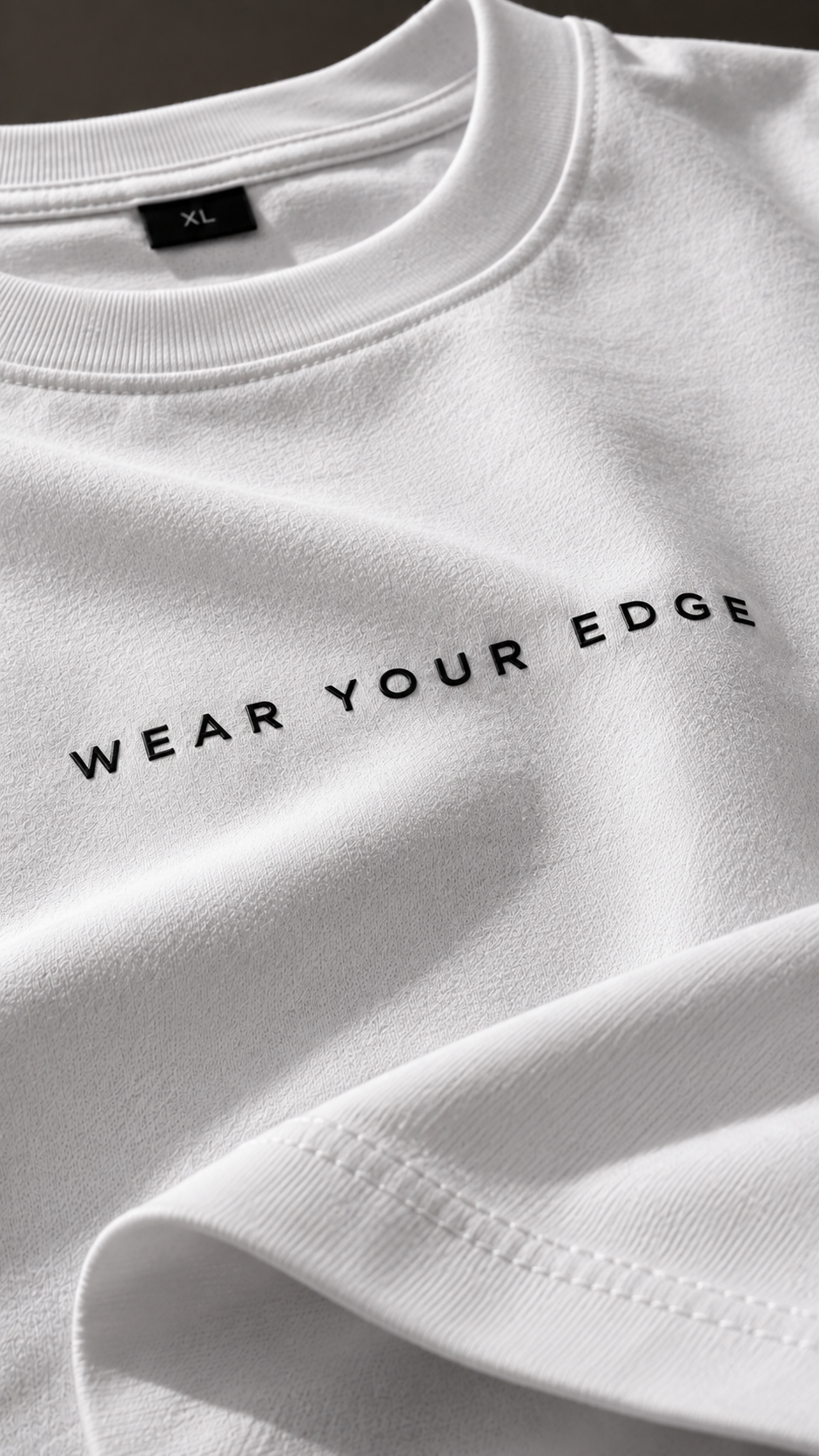 A close-up of a white crewneck t-shirt with the black text 'WEAR YOUR EDGE' printed across the chest. The fabric is shown with soft folds, and a black neck label reading 'XL' is visible inside the collar.