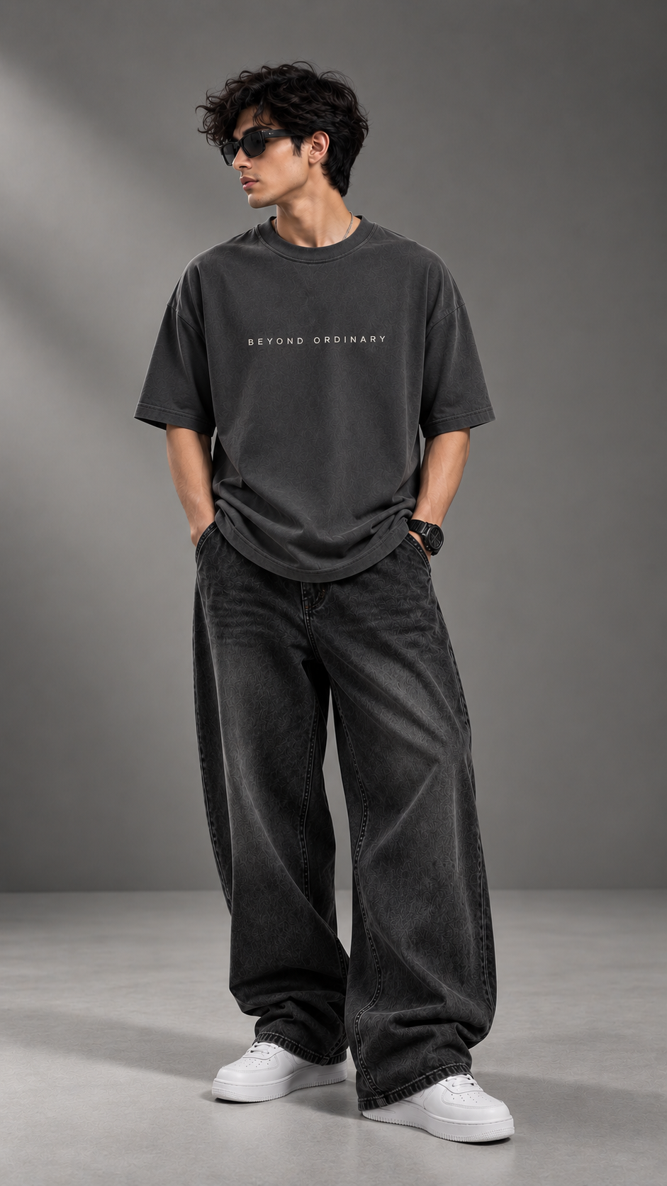 A man wearing a dark charcoal gray oversized t-shirt with 'BEYOND ORDINARY' printed on the chest, paired with matching dark gray baggy jeans and white sneakers, standing against a gray studio background.
