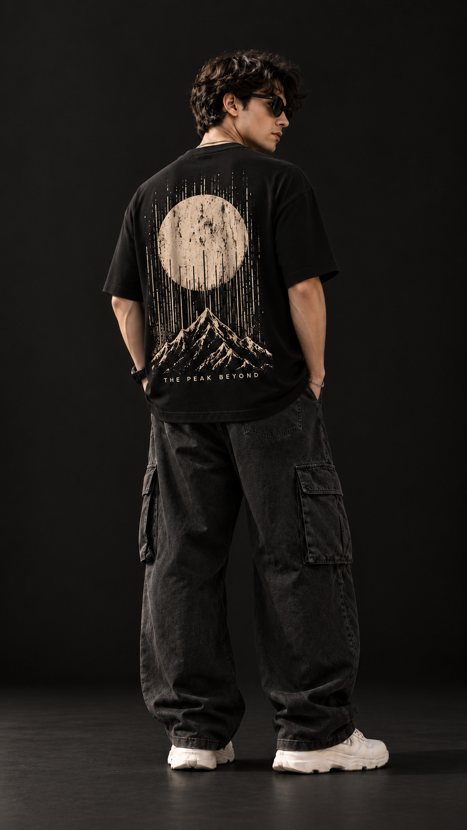 A man wearing a black graphic t-shirt with a large cream-colored mountain and moon design on the back, paired with black cargo pants and white sneakers, photographed against a dark studio background.