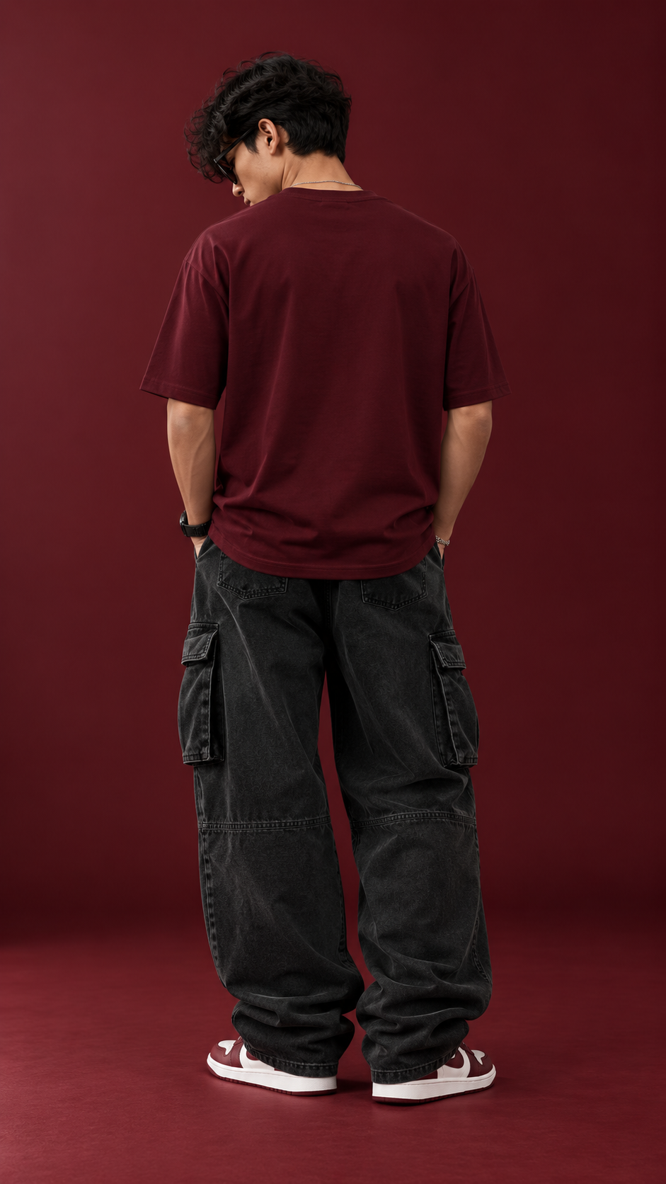 A man viewed from behind wearing a dark burgundy t-shirt, black cargo pants, and red and white sneakers, standing against a solid dark red studio backdrop.