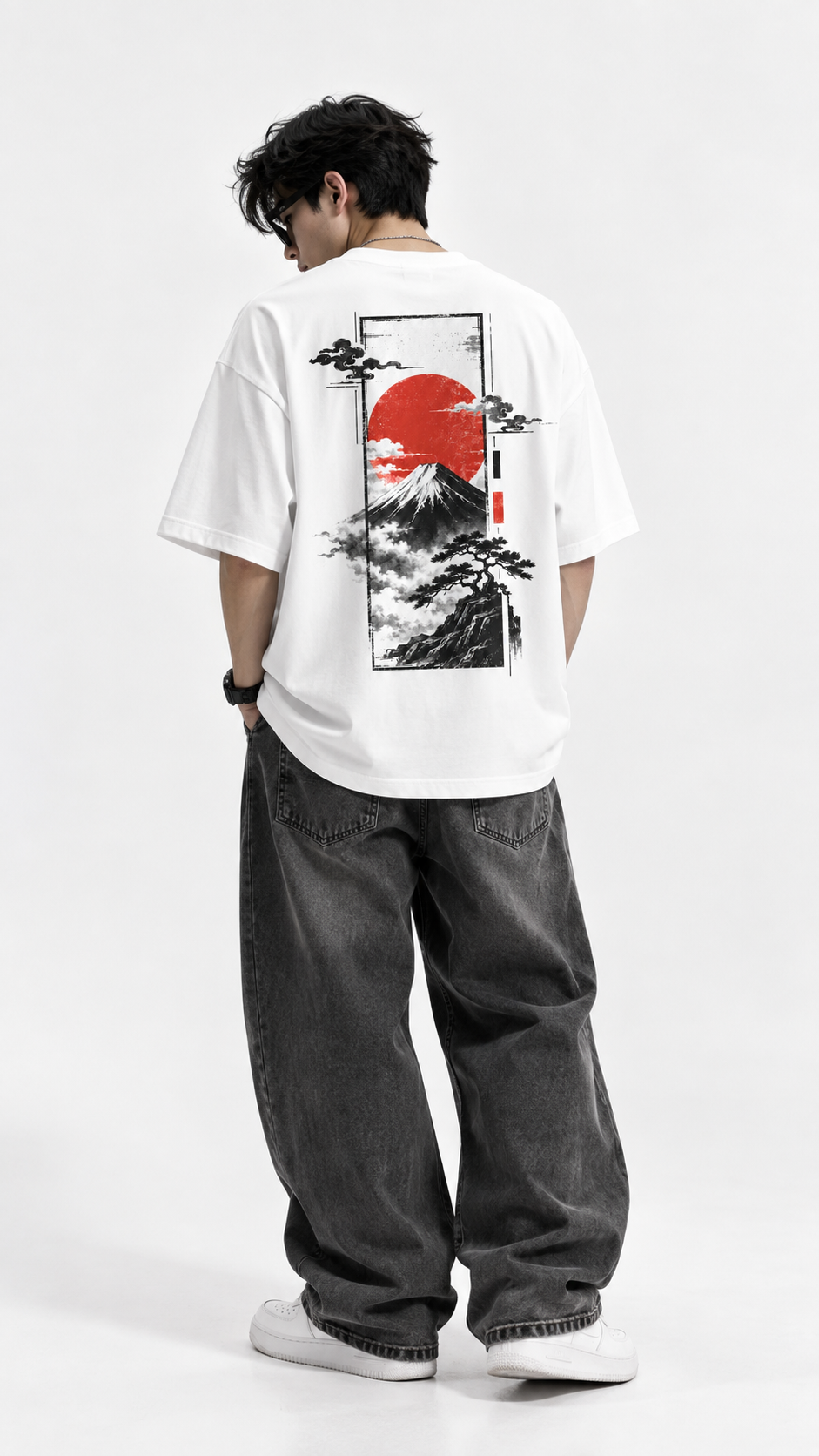 A man viewed from behind wearing a white oversized t-shirt with a large Japanese-style graphic print on the back, featuring a red sun, black mountains, and clouds. He is also wearing loose dark gray jeans and white sneakers against a plain white studio background.