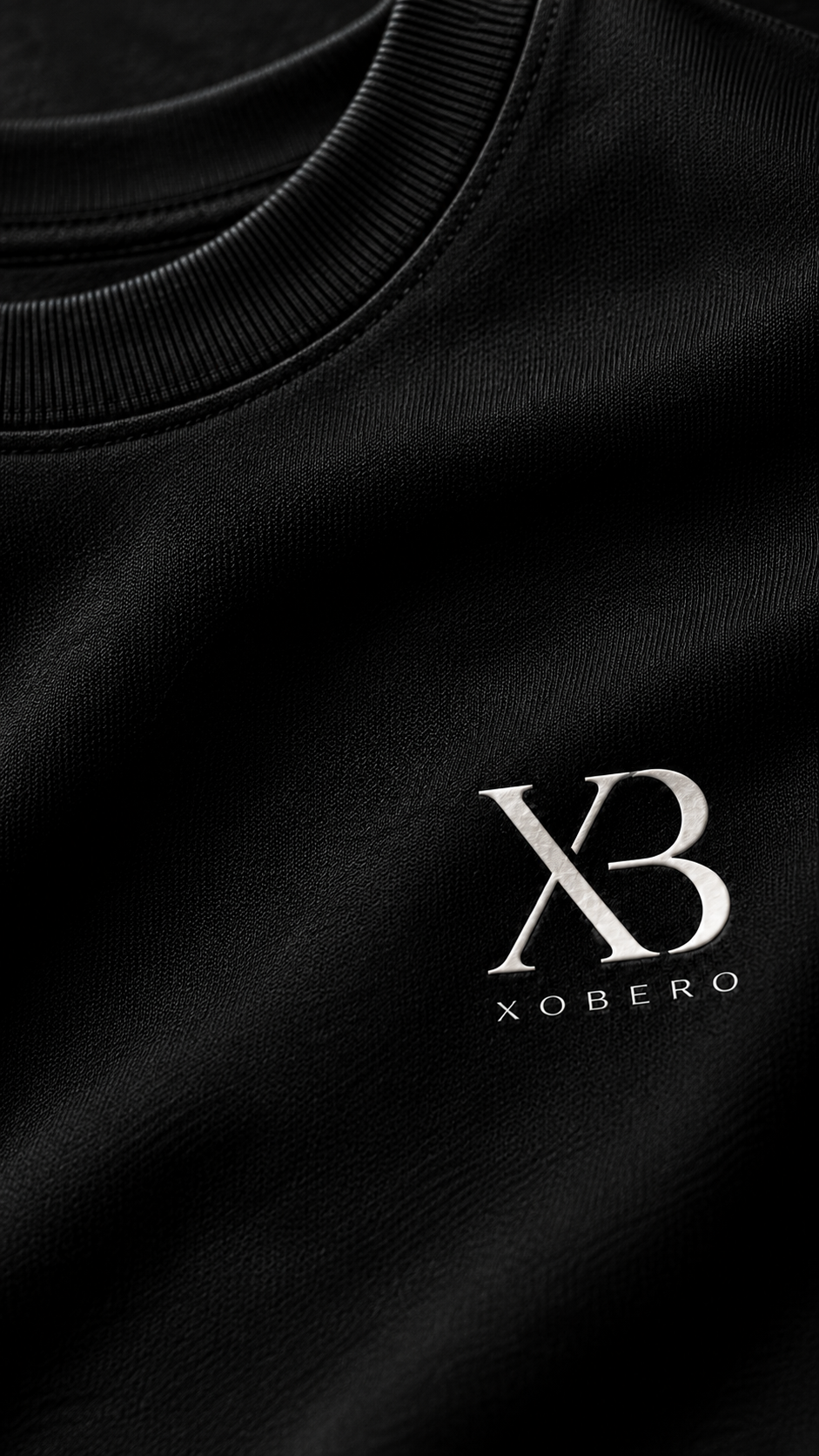 Close-up of a black crewneck sweatshirt featuring a silver metallic 'XB XOBERO' logo on the left chest.
