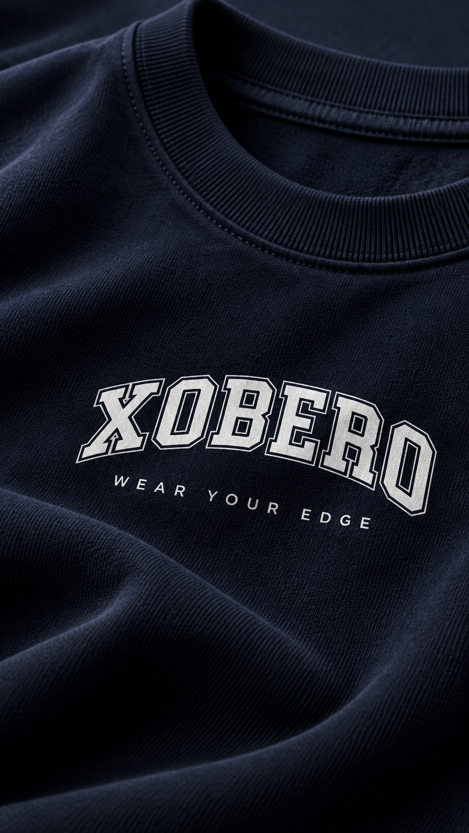 Close-up of a dark navy crewneck sweatshirt featuring the word 'XOBERO' in white varsity-style lettering with a black outline, and the phrase 'WEAR YOUR EDGE' printed underneath.